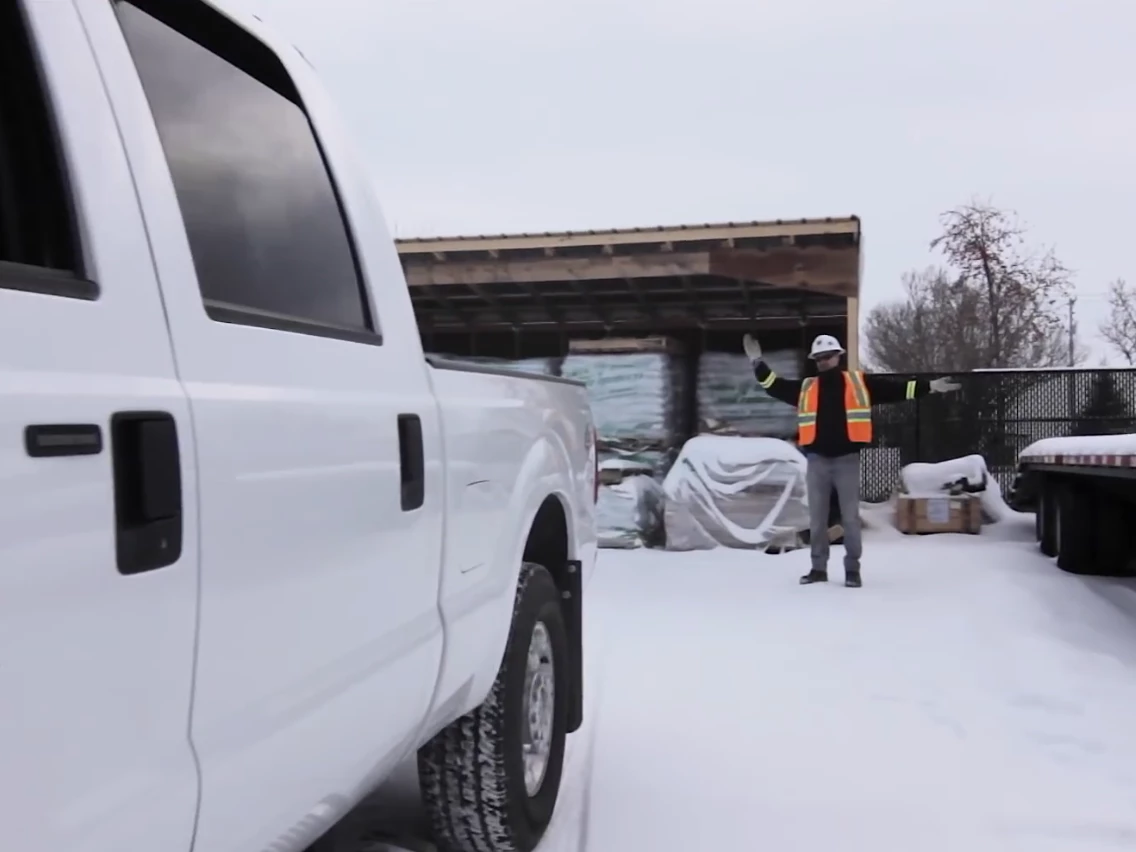 A safety-certified spotter in a hard hat and high-visibility vest directs a large white pickup truck in a snowy industrial yard.