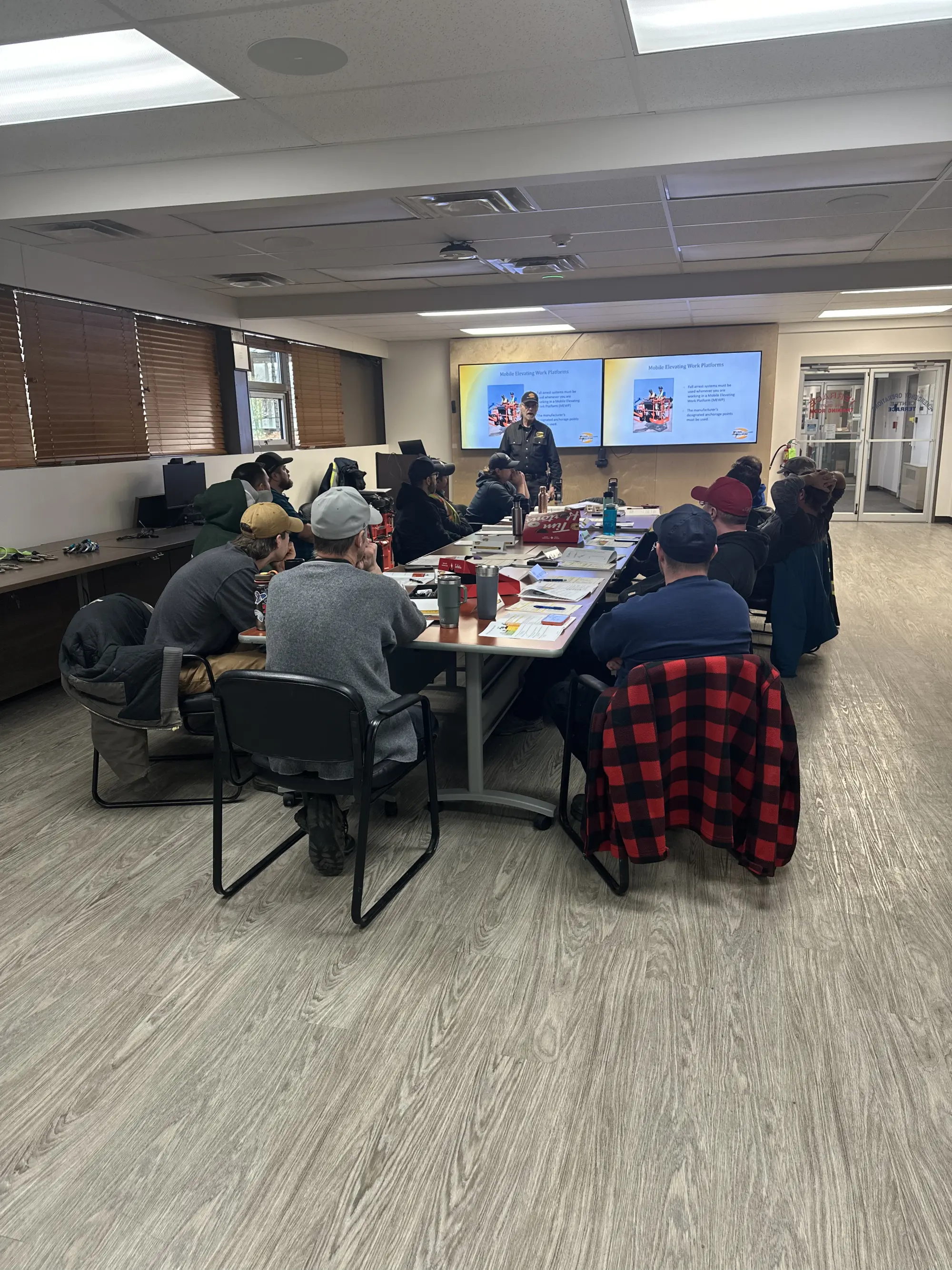 A group of students attentively listening to an instructor during a workplace safety training session conducted by Karma Training in Northwestern BC. The instructor is presenting information on mobile elevating work platforms on a large screen. This hands-on training program adheres to WorkSafeBC regulations.